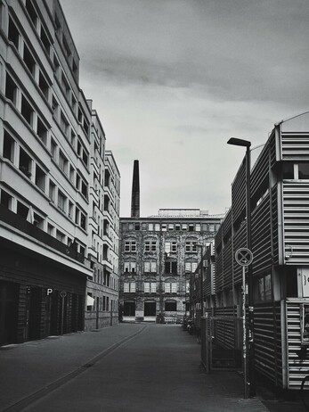 A black and white, eye-level, full shot shows a street view looking toward a tall, slender chimney on a large building, positioned centrally in the background. The street is flanked on the left by a multi-story building with numerous windows, and on the right by a shorter, modern-looking building with horizontal slats. A 'P' for parking sign is visible on the left building. The sky is overcast.