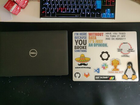 The image shows a workspace with a closed Dell laptop on the left and a sticker-covered laptop on the right. The Dell laptop is black with a textured surface and the Dell logo prominently displayed in the center. The sticker-covered laptop has a white sticker with various texts and graphics, including "I'M HERE BECAUSE YOU BROKE SOMETHING," "WITHOUT DATA IT'S JUST AN OPINION," and "HAVE YOU TRIED TO TURN IT OFF AND ON AGAIN??" The sticker also features a sombrero, a gear, a penguin, and logos for various software and hardware, such as Apple, Microsoft, and Linux. Above the sticker-covered laptop, there is a mechanical keyboard with backlit keys, some of which are blue, and a red box with white text in the top left corner. The desk surface is dark, and there are some small debris scattered around the laptop.
