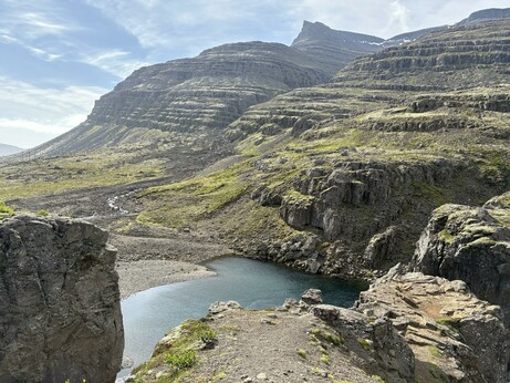 Mountains with many layers of rock running at a slight diagonal across the picture. There is a small lake in the foreground and blue skies with a few clouds above