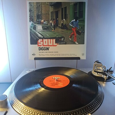 A vinyl record sits on a turntable. Behind the turntable, a vinyl album outer sleeve is displayed. The front cover shows people walking down and hanging out on a city street. There is a parked car with a damaged hood.