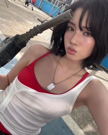 The image features a young woman, Kpop idol Yoon, wearing a red tank top under a white sleeveless top. One unfastened shoulder strap from the white top has come down to hang beneath her bust. She wears  a necklace with a dog tag pendant and is holding a petrol pump  hose and nozzle,  with the metal tip of the nozzle positioned near her head. The background shows a concrete surface with blue and white markings, and there are some elements, such as a blue fence and a red object in the distance, that could be found at a garage or petrol station. Yoon's hair is short and dark, and she is looking directly at the camera. The lighting suggests it is daytime.

Provided by @altbot, edited by @jemmesedi