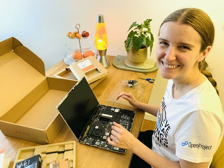 A woman is working on a laptop on a wooden table, with its screen open and components exposed. She is smiling and wearing a t-shirt with the OpenProject logo. Nearby, a brown cardboard box is open.