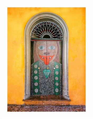 This photography is of a vividly coloured, arched window adorned with an ornate, surreal painting. The artwork depicts a face with wide, blue eyes, a protruding tongue, and a diamond necklace. The subject is an animal with human-like features. (I think it's a cat but I'm not too sure) The background is an orange wall, and the door frame is made of stone and gray. The painting style is reminiscent of folk art due to all the intricate line work (but I mean, I'm not an art doctor)