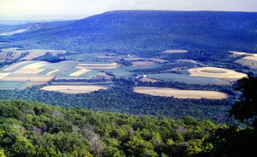 We are standing at an outlook along the long flat crest of a mountain looking towards a similar wooded mountain a few miles away. The wooded slopes of our mountain run down towards a valley between the two mountains. The flattish valley floor is mostly green and golden brown farm fields, with a few narrow strips of forest as wind breaks and a handful of visible farm buildings. Through a gap in the other mountain on the left of our picture, we can just make out a similar farming valley in the hazy distance with another line of mountains beyond. The sky is bright and sunny, but very hazy and whitewashed.