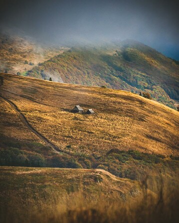 Połonina Wetlińska in Bieszczady Mountains, Poland – panoramic view of golden grassy slopes with two rustic wooden huts under cloudy skies. A hiking trail winds through the scenic highlands, surrounded by lush forests and misty ridgelines in this remote Eastern Carpathian landscape.