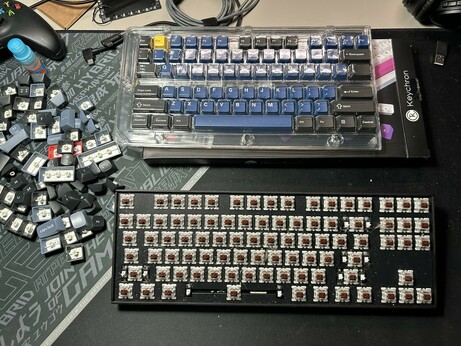 Image of my desk with a pile of the original keycaps to the left, a "naked" keyboard with only brown switches seen, and the new keycpas in the plastic storage above it.