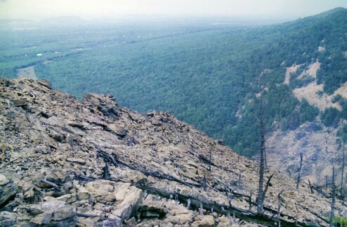 We are standing hundreds of feet up a mountainside looking down at a river valley ravaged by industrial air, ground and water pollution. Beneath a whitewashed hazy sky the AT climbs through a surreal wasted landscape of naked rock — gigantic boulders covered with chunks of blasted rubble. Occasional black, broken skeletons of dead trees grope upward through the barren mass. There are continuous panoramic views into the smoky industrial valley below. Beyond the heavily localized devastation, the valley and the mountainside across the river from us is heavily wooded. Distant views are heavily obscured by the steamy, dirty air.