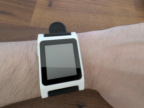 A picture of my wrist against a dark brown desk.  On my wrist is a white rePebble Core 2 Duo smart watch on a black strap.  It's big and stretches the width of my wrist.  The screen is inlaid with a black bezel.  The screen is turned off.
