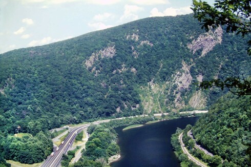 Beneath a light blue sky with puffy clouds a dark blue river makes a gentle S curve through a deep gorge between two mountains. The mountainside on the right is heavily wooded and has a railroad and a two-lane road running along its base beside the river. The second mountain forms the background of the picture. It is tall and the side facing us looks almost vertical, with several naked rock cliffs among the trees. A 4-lane interstate highway runs along the river bank at its base.