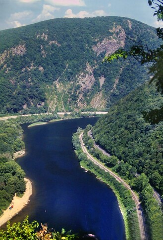 Beneath a light blue sky with puffy clouds a dark blue river makes a gentle S curve through a deep gorge between two mountains. The mountainside on the right is heavily wooded and has a railroad and a two-lane road running along its base beside the river. The second mountain forms the background of the picture. It is tall and the side facing us looks almost vertical, with several naked rock cliffs among the trees. Another road runs along the riverbank at its base.