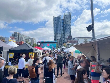 Eine geschäftige Marktszene des Veganen Straßenfestes in Hamburg im Freien mit zahlreichen Zelten und Ständen. Menschenmengen versammeln sich und beschäftigen sich mit Anbietern. Im Hintergrund zeigt eine große Leinwand ein Ereignis, eingerahmt von modernen Gebäuden unter einem teilweise bewölkten Himmel.