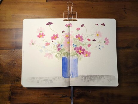 This is a watercolor painting of summer wildflowers in a blue ceramic vase. It's painted across the spread of two pages in a B5 portrait sketchbook.  It's mostly cosmos in various shades of pink with yellow centers, peach allium, dark red scabiosa, and light blue larkspur.