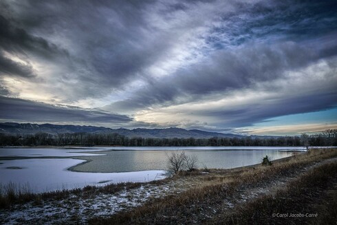 A partially frozen reservoir reflects the stormy skies above it just shortly before the sun sets.  The ice on the water is covered with a layer of snow, but the shoreline of grasses and trees has almost no snow. The mountains in the distance are hazy and gray, mostly just a silhouette.  The cloud layer above the mountains is dense, dark gray, and it looks stormy and blocks any view of the setting sun. Another layer, interspersed with stripes of blue sky, looks light and fluffy, blown by the wind into long light gray and cream stripes running from west to east. There is a bright blue patch of sky to the north, and yet another distant pale layer of clouds.