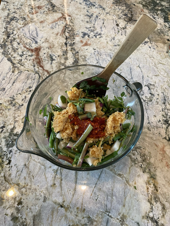 The clear batter bowl  is seen from above. We see large slices of purslane in green and purple, cubes of daikon in white and purple, Cherokee green beans, golden garlic paste, shiny red chili paste, brown ginger paste.green