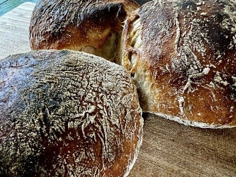 Auf einem dunklen Holztisch liegt ein Holzbrett. Darauf liegen zahlreiche frisch gebackene Brötchen mit rustikalem Ausbund