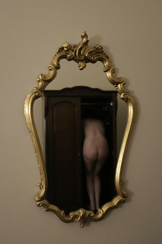 Ornate gold-framed wall mirror reflecting a person entering a wardrobe, visible from the back. The room has beige walls, and the wardrobe has a dark wooden finish with a closed door on one side.