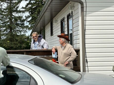 Two people outside of a house. There is an older woman, Marion Knudsen along with an older man, Tako, who is leading the field trip. There is a car in the foreground.