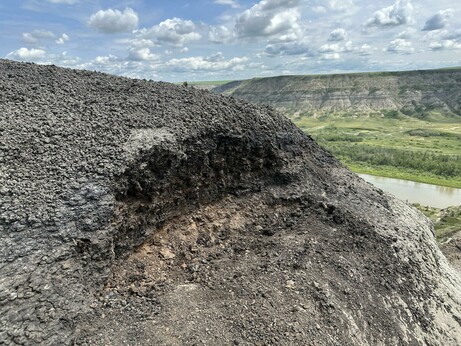 The side of a hill with a small excavation. In the excavation you can see the colour change from brown / black to orange; that's the K-Pg extinction layer. In the background you can see the badlands and the river running through them.
