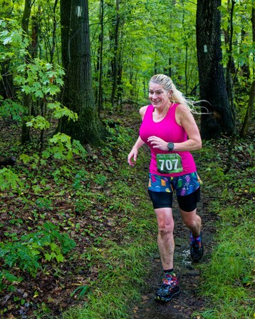 A woman with blond hair and a pink tank top has a green bib that say 707 in white is running on a path between two trees with rectangular white blazes