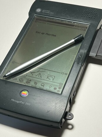An Apple Newton MessagePad 2100 is dark gray plastic with a stylus on top of the screen. A 3D printed battery tray is peaking out of the bottom left. On the screen it says "Eat up Martha"