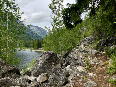 A rocky trail on the far right passes above large blocks below, forest ahead. On the left and below is a lake, a mountain rising from its shore, another mountain in the distance. The sky is cloudy.