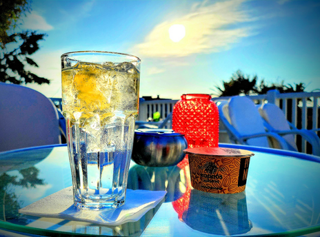 Auf einem Cafetisch auf einer Terrasse mit Loungemöbeln steht ein Cocktailglas gefüllt mit Eis, Zitronenscheibe und einer durchsichtigen Flüssigkeit (Gin und Tonic). Unbedeutend daneben ein glänzend leerer Aschenbecher, ein Bercherchen Nüsse und ein rotes Teelichtglas.
