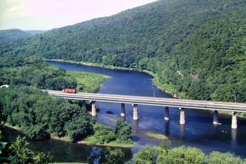 Beneath a pale blue sky with hazy clouds, a wide river forks around several large wooded islands. A wooded mountainside rises steeply from the far riverbank. A long multi-span beam bridge supported by concrete piers carries a 4-lane interstate from the far riverbank on our right to one of the islands. Traffic is light, only 2 trucks and 1 car are visible.