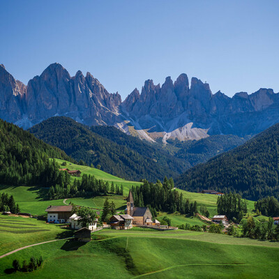 Chiesa di Santa Maddalena in Val di Funes, South Tyrol, Italy, perched on a grassy slope with its pointed steeple framed by the dramatic Odle mountains, surrounded by alpine cottages and sunlit meadows under a vivid blue sky.