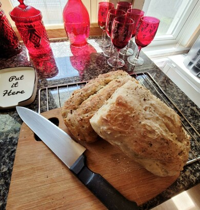 This image captures a cozy kitchen scene with a fresh loaf of homemade Italian bread being sliced on a wooden cutting board. The bread is rustic and golden brown with a textured, herb-speckled crust, and a few slices have been cut to reveal a soft, fluffy interior. A sharp kitchen knife with a black handle lies next to the bread.

The countertop is a dark, glossy granite, and sunlight pours in through a nearby window, casting a warm glow. To the side, there’s a wire cooling rack beneath the bread. On the counter is a small white dish with black text that reads “Put it Here.”

Lining the windowsill and counter are several decorative red West Virginia glass items: a tall, patterned jar with a lid, a bottle, and a row of vintage-style goblets with floral etchings, all in deep ruby red, giving the scene a charming, nostalgic touch. The overall atmosphere is inviting, homey, and full of warmth.