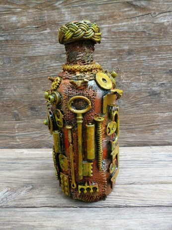 This image features a bottle that has been transformed into a compelling piece of steampunk-style art. Dominating the scene, the bottle is intricately adorned with an assortment of small mechanical components—gears, springs, bolts, and screws—all arranged with meticulous care. These metallic elements, some brass-colored and others with a more muted, rust-toned patina, create a textured and tactile surface that invites closer inspection.

The bottle itself shows a warm, earthy base color beneath the metallic embellishments, blending hues of orange, brown, and copper. This color palette enhances the antique and industrial vibe typical of the steampunk aesthetic. The bottleneck is wrapped in a thick, woven cord that adds a rustic yet deliberate finish to the piece, giving it a handcrafted feel. The backdrop, a rough wooden surface with visible grain, contrasts with and highlights the bottle's mechanical intricacies, grounding the artwork in a natural, raw environment.

Artistically, this creation evokes themes of invention and the fusion of organic and mechanical life, hallmarks of the steampunk genre, which melds 19th-century industrial steam-powered machinery with fantasy or speculative technology. Symbolically, this transformation of a mundane bottle into a miniature machine-like relic could suggest ideas about recycling, the beauty of repurposing objects, or the potential for hidden complexity within the everyday.
