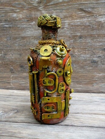 This image features a bottle that has been transformed into a compelling piece of steampunk-style art. Dominating the scene, the bottle is intricately adorned with an assortment of small mechanical components—gears, springs, bolts, and screws—all arranged with meticulous care. These metallic elements, some brass-colored and others with a more muted, rust-toned patina, create a textured and tactile surface that invites closer inspection.

The bottle itself shows a warm, earthy base color beneath the metallic embellishments, blending hues of orange, brown, and copper. This color palette enhances the antique and industrial vibe typical of the steampunk aesthetic. The bottleneck is wrapped in a thick, woven cord that adds a rustic yet deliberate finish to the piece, giving it a handcrafted feel. The backdrop, a rough wooden surface with visible grain, contrasts with and highlights the bottle's mechanical intricacies, grounding the artwork in a natural, raw environment.

Artistically, this creation evokes themes of invention and the fusion of organic and mechanical life, hallmarks of the steampunk genre, which melds 19th-century industrial steam-powered machinery with fantasy or speculative technology. Symbolically, this transformation of a mundane bottle into a miniature machine-like relic could suggest ideas about recycling, the beauty of repurposing objects, or the potential for hidden complexity within the everyday.