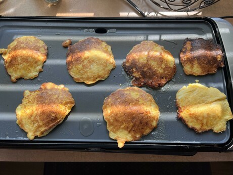 Photo of seven yellowish pancake shaped objects on a flat cooking griddle.