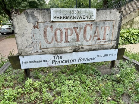 A weathered sign for "CopyCat of Evanston" is displayed, featuring a cartoon cat character. Below it, there’s a promotional section for "The Princeton Review" with contact information. Lush greenery and a pathway are visible in the background