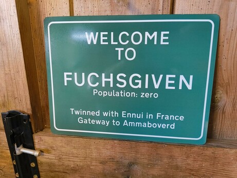 A green metal sign with white text in the style of British road signs. The test reads "welcome to Fuchsgiven, population zero" Beneath is a subtext reading "twinned with Ennui in France, gateway to Ammaboverd"