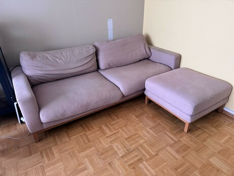 as Bild zeigt ein modernes, lila Sofa mit einem angrenzenden Ottomana in einem Raum mit hellen Wänden und einem parquetbemusterten Holzboden. Das Sofa hat eine L-Form und besteht aus weichen, flauschigen Polstern, die in einem einheitlichen lila Ton gehalten sind. Die Armlehnen sind breit und ergonomisch gestaltet, und die Rückenlehnen sind leicht gepolstert. Der Ottomana ist ebenfalls in lila gehalten und bietet zusätzlichen Platz für die Füße oder als zusätzliche Sitzfläche. Die Beine des Sofas und des Ottomanas sind aus Holz und haben eine natürliche Farbe, die gut zum Holzboden passt.