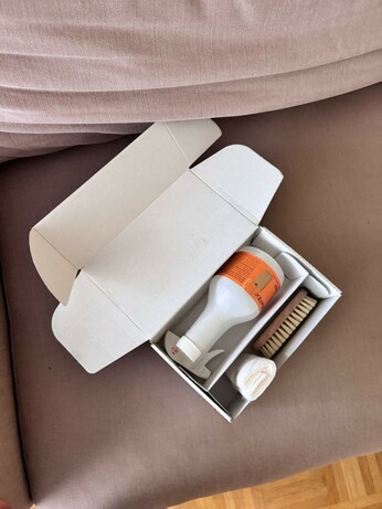 Das Bild zeigt eine offene weiße Kartonbox, die auf einem altrosa Sofa liegt. In der Box befinden sich drei Gegenstände: ein weißes Flaschenprodukt mit einem orangefarbenen Etikett, ein Bürstenset mit einem Holzgriff und weichen Büscheln, sowie ein weißes Tuch. Die Bürste ist in einer speziellen Nische der Box platziert, während das Tuch zusammengerollt ist. Die Box selbst ist einfach gestaltet und hat eine klare, funktionale Struktur. Der Hintergrund besteht aus einem beigen Sofa, der die Gegenstände gut zur Geltung bringt.