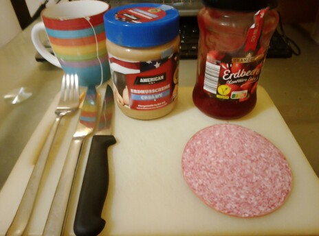 A fresh cuppa tea steeps, and fixin's for sweet and sour pork sandwiches, creamy peanut butter, strawberry preserves, and salami, along with cutlery, sit on a white acrylic cutting board.