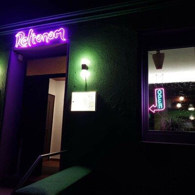 Eine nächtliche Ansicht der aussenfassade unserer Bar, mit leuchtendem Retronom-Schild, Lampen und einem Neon "Open" Schild.