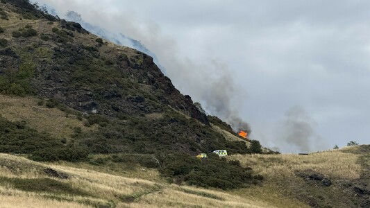 Arthur’s Seat, an extinct volcano in Edinburgh. A fire is burning on the west side of the hill. A couple of police cars are parked on the road and a fire burns behind them.
