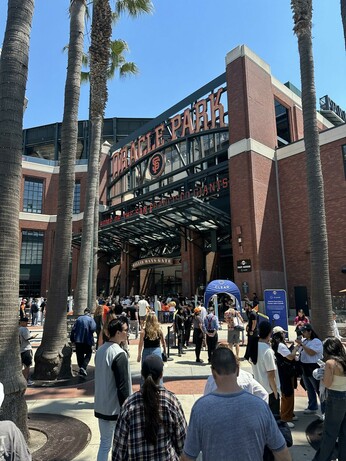 The Willie Mays gate, entrance to Oracle Park, a baseball stadium. It’s basically an opening with brick walls on the sides and overhead neon block lettering reading “Oracle Park” and “Home of the San Francisco Giants” and the block lettered “SF” logo. The plaza in front of the entrance is moderately crowded and there are several palm trees in the foreground.