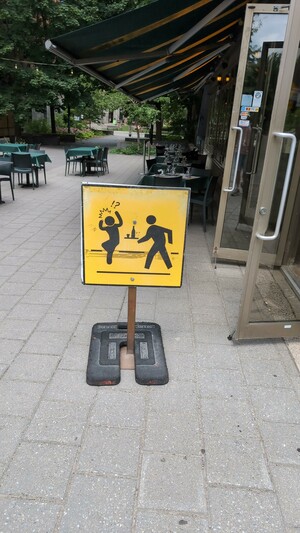 A square, yellow sign with a person walking to someone dancing and surprised, with a bar table (with drinks) behind them. The sign is standing from the sidewalk.