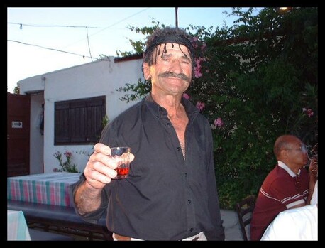 Middle-aged man with distinctive moustache and headband holding a small glass of red drink at an outdoor evening party in a Cretan village, with white buildings and flowering trees in the background.