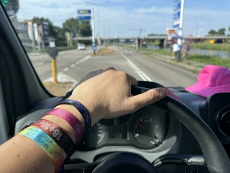 A steering wheel held by a hand with 4 WHY wristbands on it. Pink WHY hat on the dashboard.