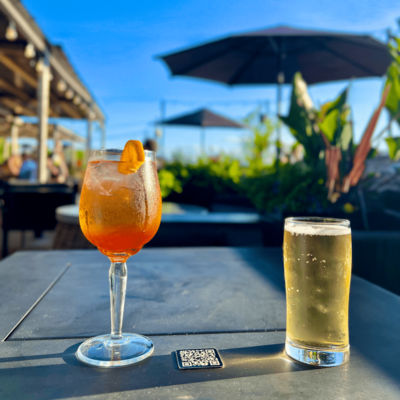 The image features a table set outdoors under a clear blue sky. On the table, there are two drinks: a large, round glass filled with a vibrant orange-red beverage, garnished with a slice of orange, and a tall, slender glass containing a light-colored beer. Both drinks are placed on a dark surface, with a QR code coaster visible beneath the orange drink. In the background, there are green plants and umbrellas, suggesting a lively outdoor setting.