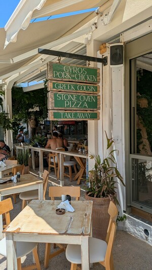 A green tavern sign on the island of Crete, listing
"Gyros – Pork or Chicken
Greek Cousin
Stone Oven Pizza
Take Away"