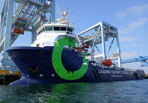 Blue offshore supply vessel with white superstructure and a large green circle painted on its side, tied up in front of two large container cranes.