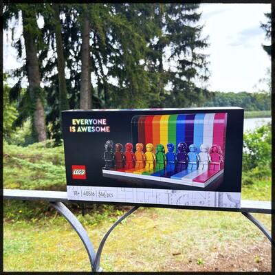 Photos of a LEGO kit package standing on a metal guardrail. The background is a garden with some trees and a lake in the background. The guardrail might be from a terrace. The LEGO kit is the "Everyone is awesome" kit which is a set of 11 figurines standing on some sort of stage. The figurines and their lane on the stage are all different colors. Together they resemble the rainbow LGBTQ+ .