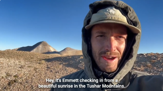 A person wearing a gray jacket stands on a treeless ridge with mountains in the background.  He has the hood of his jacket pulled up over a baseball cap, so all you can see is his face as he introduces himself and describes the view of the surrounding landscape.