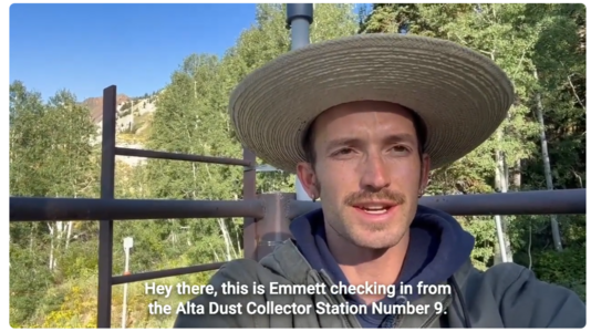 A person wearing a hat with a wide brim for sun protection speaks to the camera with green trees and blue sky in the background.