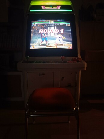 Un cabinato arcade in penombra, illuminato solo dal tubo catodico e dalle luci nella parte superiore. Sul monitor c'è la schermata di un gioco di combattimento.